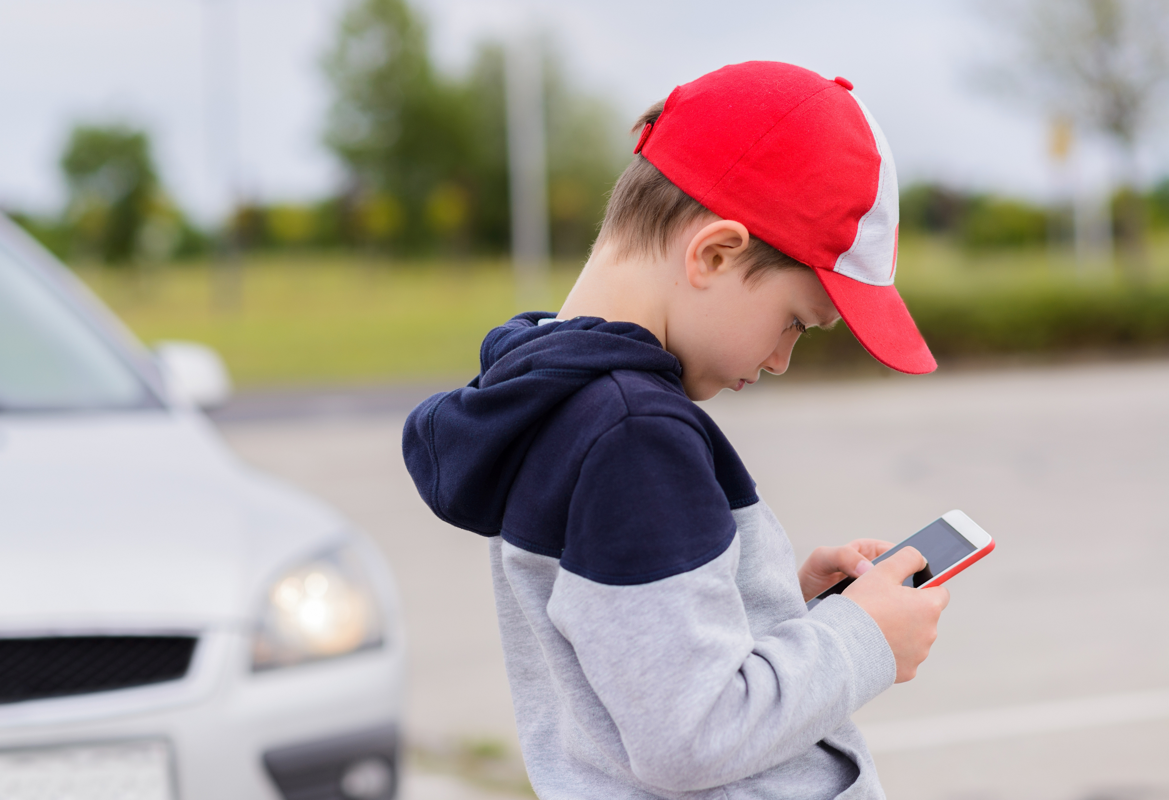 Gutt med caps stirrer intenst på mobiltelefonen. En bil nærmer seg i bakgrunnen uten at han tilsynelatende legger merke til det.